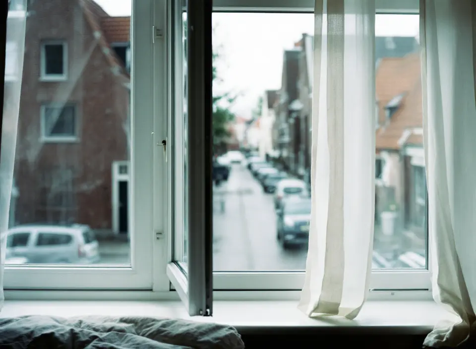 Slaapkamer met een open raam en een vitrage die beweegt in de wind, met bakstenen huizen aan de overkant